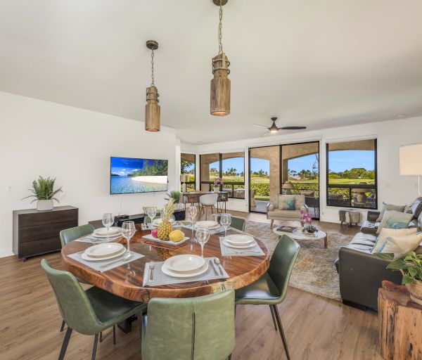 A bright, open living-dining space with a round wooden table set for six, green chairs, a sofa, TV, sunny bay windows, and plants.