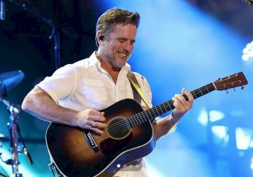 A smiling guitarist plays an acoustic on stage under blue lights, wearing a white shirt and enjoying the moment.