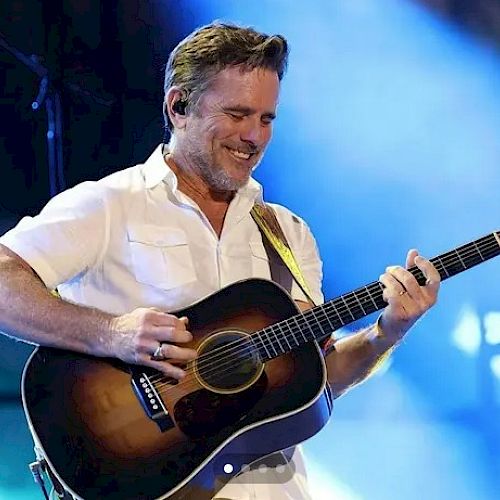 A smiling guitarist plays an acoustic on stage under blue lights, wearing a white shirt and enjoying the moment.