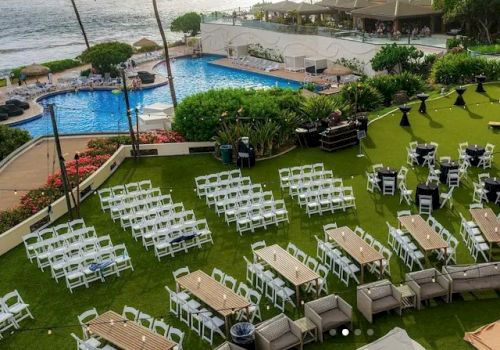 Beachside event setup with white chairs and tables on a lawn, pool and ocean view in background; guests may gather by the water.