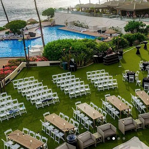 Beachside event setup with white chairs and tables on a lawn, pool and ocean view in background; guests may gather by the water.