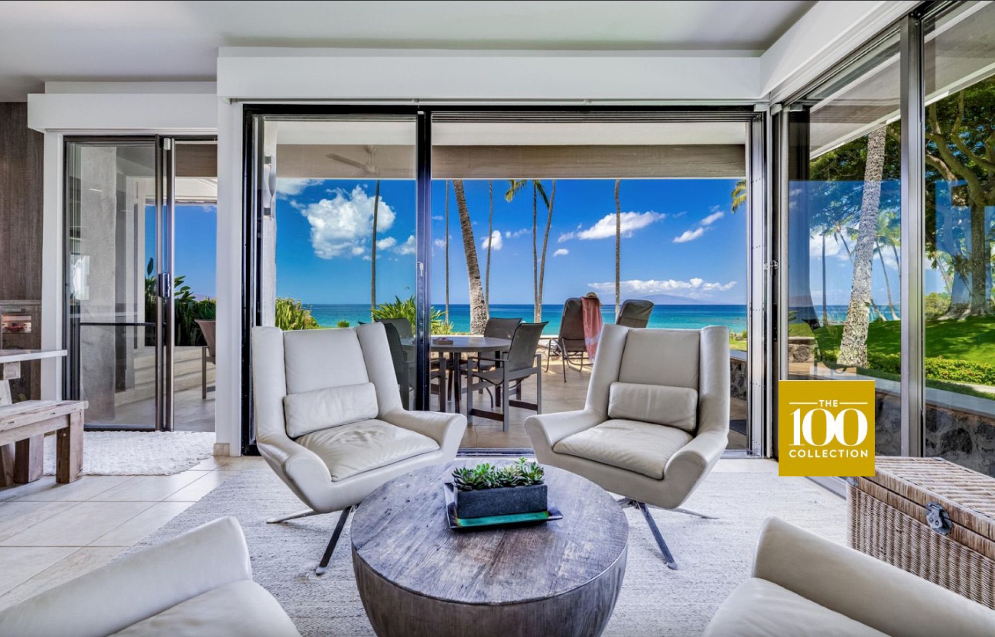 A luxurious beachfront living room with white leather chairs, a round coffee table, and glass doors opening to a patio and blue ocean view, bright and inviting.