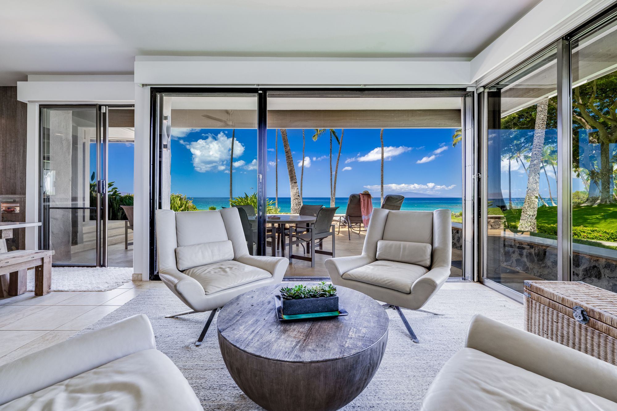 A modern open-concept living room with white leather seating, a round stone coffee table, sliding glass doors, and a sunny ocean-view patio.