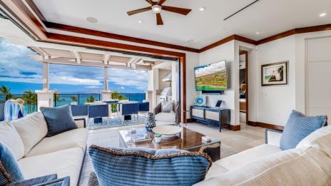 Luxurious open living area with cream sofas, blue cushions, a glass coffee table, and a balcony view of the ocean and blue sky.