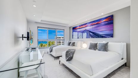 A bright, modern hotel room with two white beds, a desk, a wall-mounted TV, and a large window with a scenic ocean view.