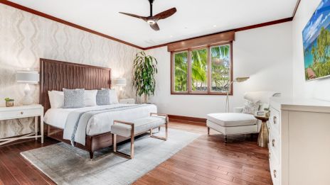 A bright, cozy bedroom with a wooden floor, queen bed, white furniture, a ceiling fan, large window, and a lounge chair by the window.