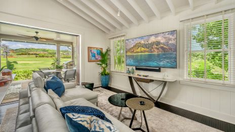 A bright, cozy living room with a large beige sectional, blue throw pillows, a wall-mounted TV, and a view into a green outdoor space through wide windows, ending here.