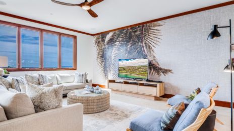 A bright, airy living room with a large beige sectional, a cozy round ottoman, palm wall mural, wood TV stand, blue lounge chair, and big windows letting in natural light, creating a tropical, relaxed vibe.