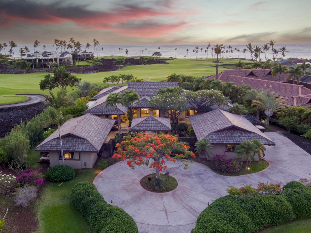 An aerial view of a tropical resort with thatch-roof villas, lush landscaping, a circular driveway, and sunset-lit sky by palm trees.
