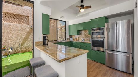Modern kitchen with green cabinets, stainless steel fridge and double oven, beige granite island, wood floor, ceiling fan, and outdoor area beyond glass doors.