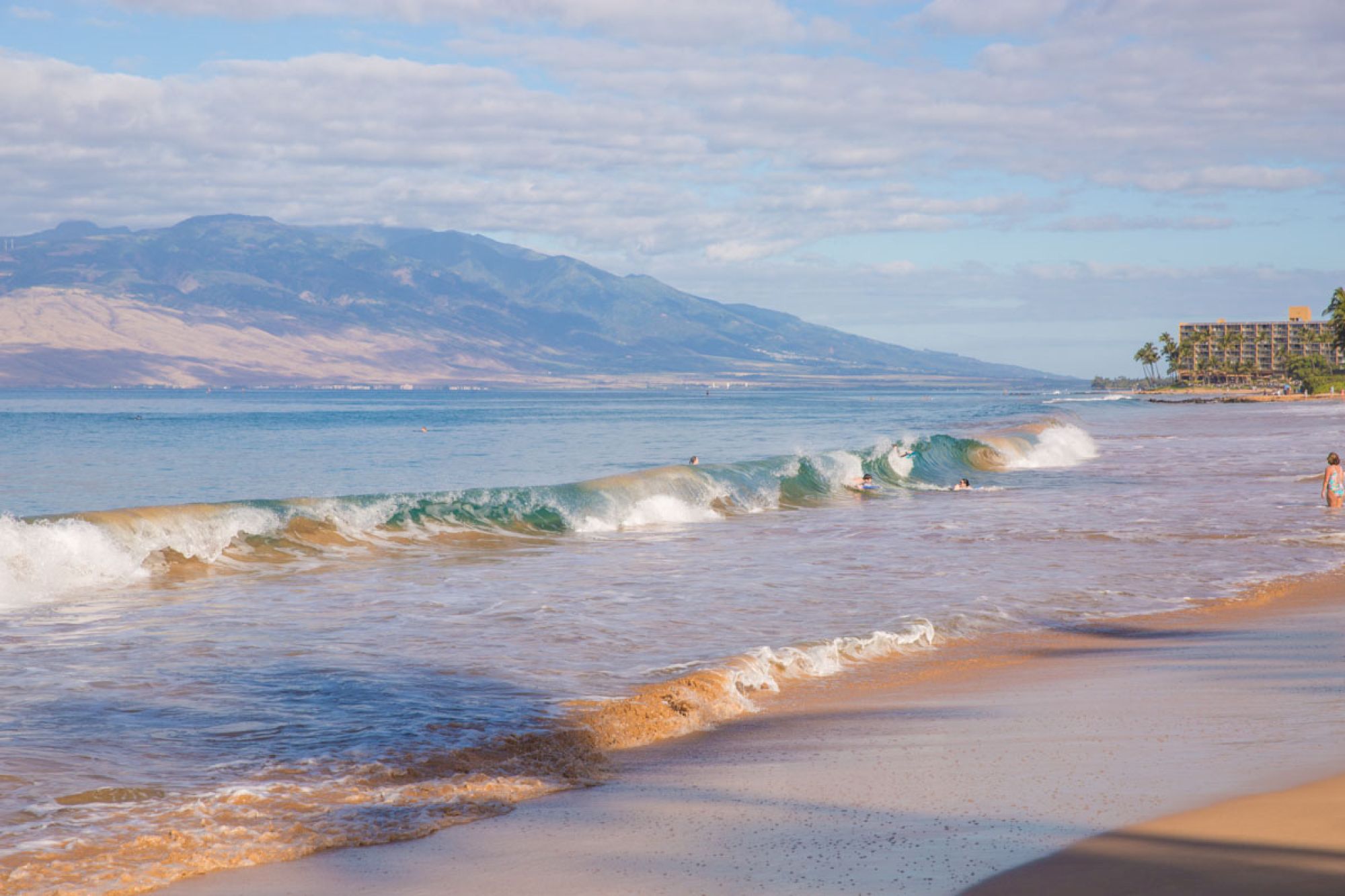 A sunny beach with gentle waves, a distant mountain range, and a few people strolling along the shoreline.