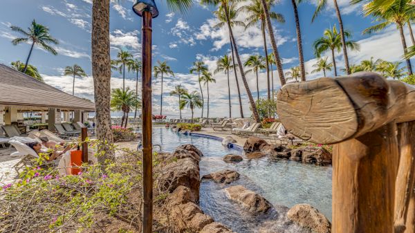 A tropical resort pool area with palm trees, stone rocks, lounging chairs, thatched roofs, and a calm sea in the background, sunny and relaxing.