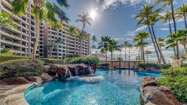 A tropical resort pool with clear blue water, surrounded by palm trees, rocks, and a sunlit beachfront hotel, perfect for a relaxing vacation.