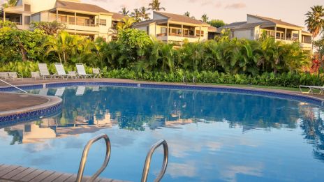 A calm hotel pool surrounded by green palm trees and lounge chairs, with pastel houses in the background, reflecting softly on the water.