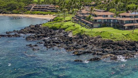 A scenic coastal resort village with turquoise water, rocky shoreline, and lush green hillsides overlooking beachfront villas.