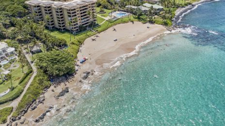 Aerial view of a coastal resort with sandy beach, turquoise water, rocky shore, and multi-story hotel buildings surrounded by greenery.