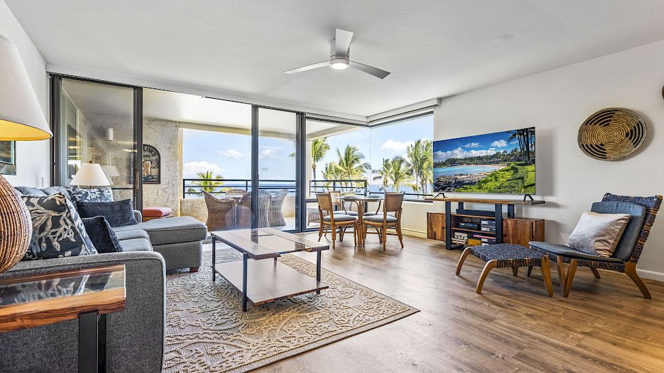 A bright, airy living room with a cozy seating area, TV, and a balcony overlooking palm trees and blue skies, perfect for relaxing indoors.