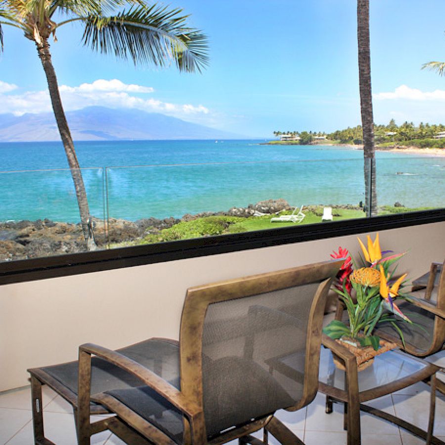 A sunny beach view through a balcony with two chairs, tropical plants, palm trees, and turquoise water beyond a low wall.