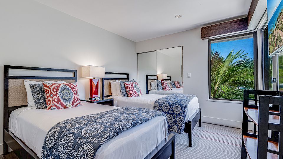A bright hotel room with two full beds, patterned blue throws, matching pillows, a nightstand with a lamp, a large mirror, and a window showing palm trees outside.