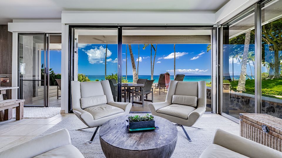 A luxurious seaside living room opens to a sunny patio with lounge chairs, a round coffee table, and sliding glass doors revealing a blue ocean view.