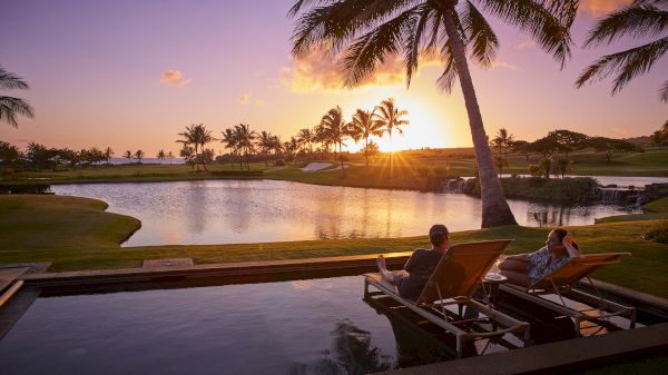 Two people lounge in wooden chairs by a tranquil pool reflecting a sunset, palm trees framing a tropical golf-like scene. Beautiful evening retreat.