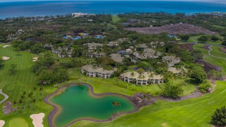 Aerial view of a lush golf course with a green pond, scattered villas, palm trees, and distant coastline under blue skies.