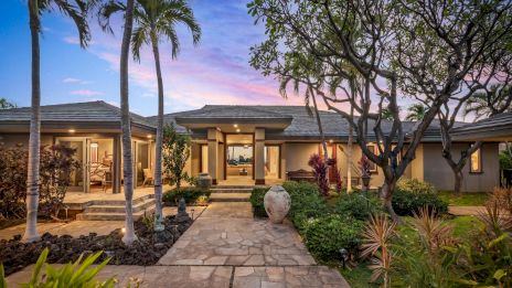A modern single-story home with a stone walkway, tropical landscaping, palm trees, and warm interior lights glowing at dusk, inviting and serene.