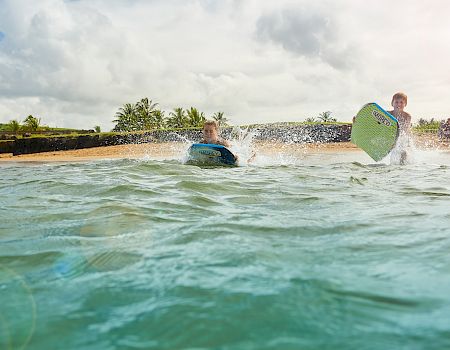 People surfing on a sunny beach: two surfers in turquoise water near a sandy shore with palm trees and onlookers; waves splash as one rides a green board.