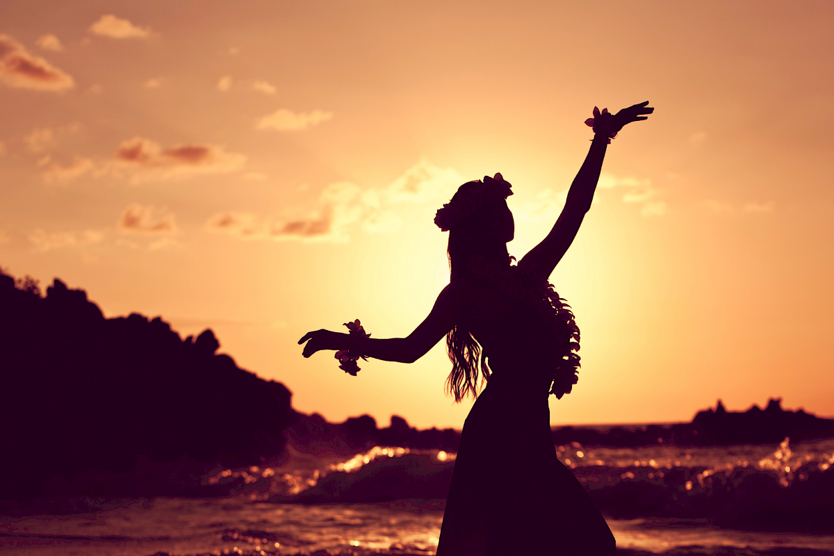 A silhouette of a person dancing on the beach at sunset, arms raised, with waves and rocky shore in the background.