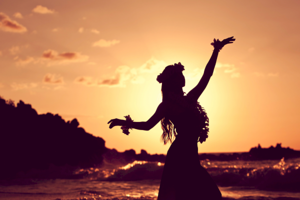 A silhouetted woman dancing on the beach at sunset, arms raised, with waves and a rocky shoreline in the golden sky.