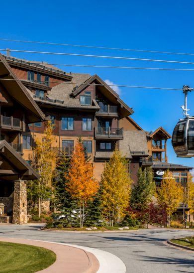 A mountain lodge with wooden architecture, autumn trees, and a gondola lift against a bright blue sky.