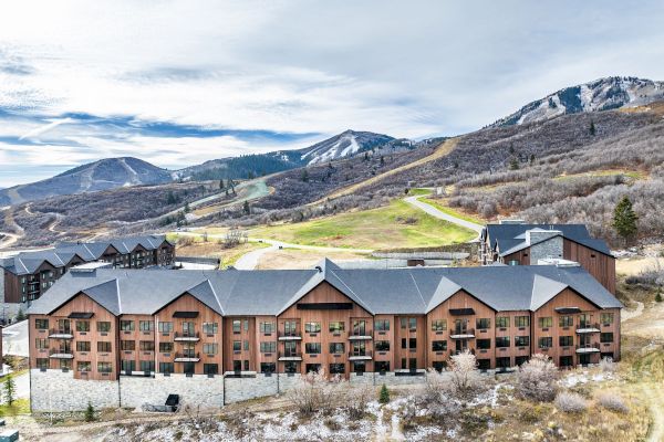 A multi-story brown brick lodge sits at the base of rolling hills, with snow-dusted mountains in the background and a winding road nearby.