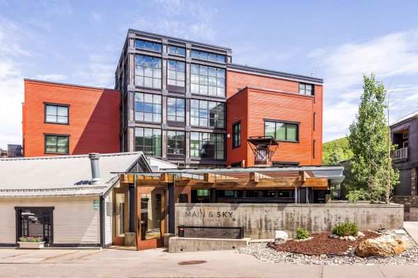 A modern mixed-use building with red siding, large glass windows, and a wood pergola entrance labeled &ldquo;MAIN & SKY,&rdquo; under a clear blue sky.
