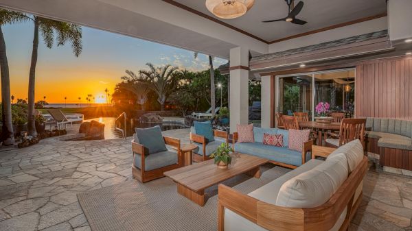 Luxurious outdoor lounge at sunset with beige cushions, wooden furniture, a stone patio, tropical plants, and a view toward the water.