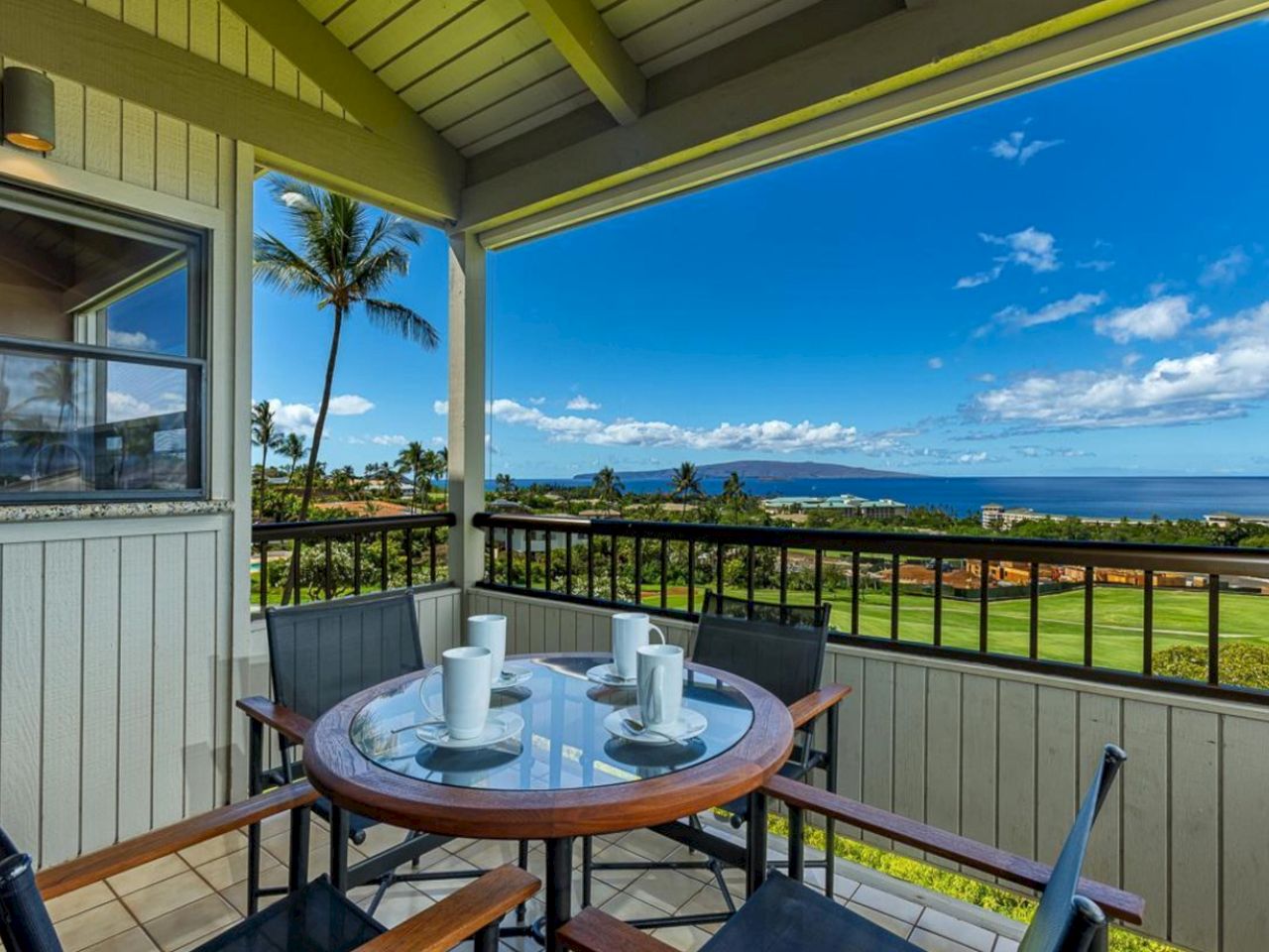 A balcony dining area with a round table set for four, overlooking a lawn and palm trees toward the ocean on a sunny day.