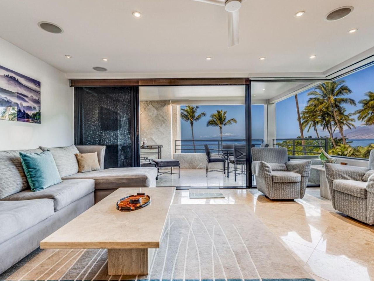 A bright, modern living room with a light gray sectional, teal accents, and a large glass door opening to a balcony with palm trees and an ocean view.