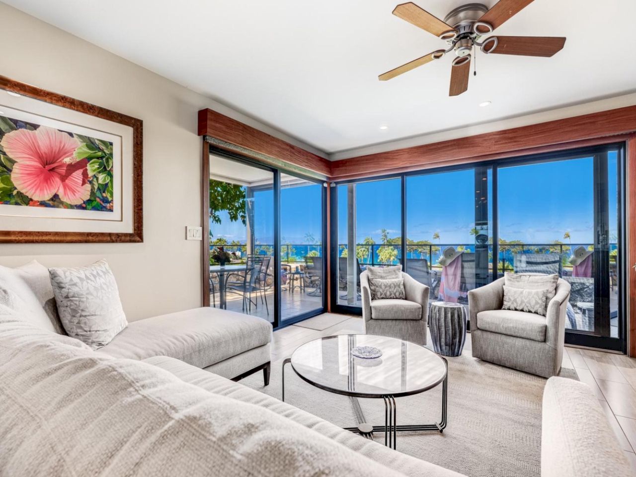 A bright coastal living room with beige sofas, a glass coffee table, and a ceiling fan; large sliding glass doors open to a balcony with ocean views.