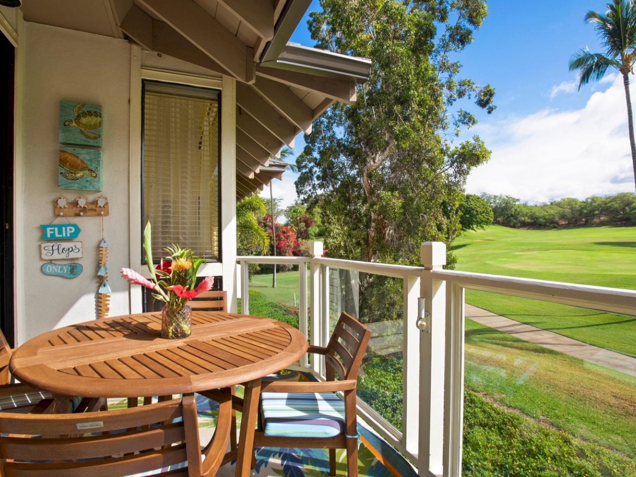A sunny balcony with a wooden round table and chairs, a flower vase, tropical plants, and a view of a green lawn and palm trees.