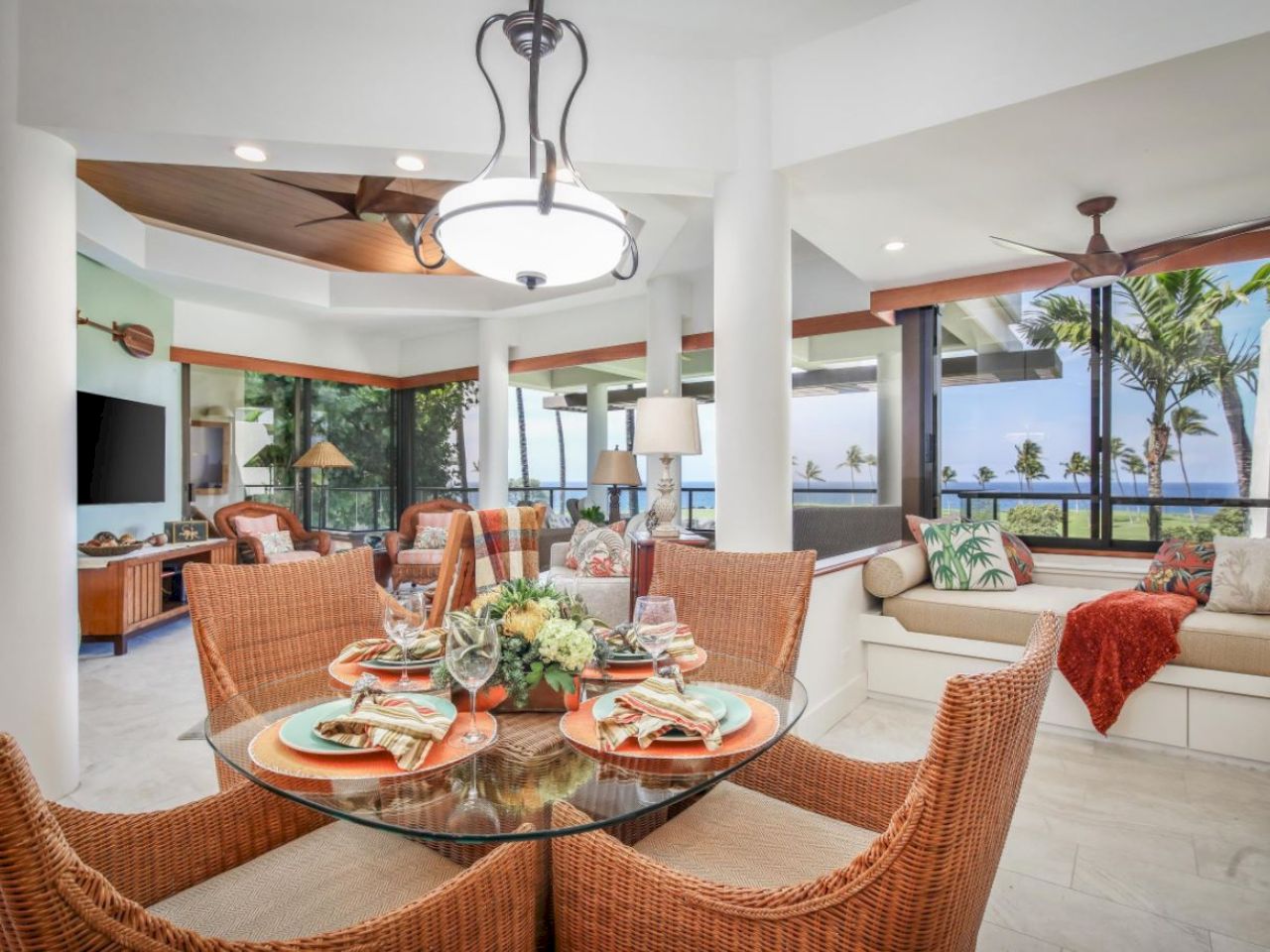 A bright tropical-style living-dining area with wicker chairs around a glass table, a beige sectional, palm plants, and large windows letting in sunlight.
