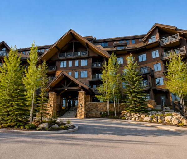 A large wooden mountain lodge with tall trees, stone accents, and a grand entrance, under a clear blue sky.