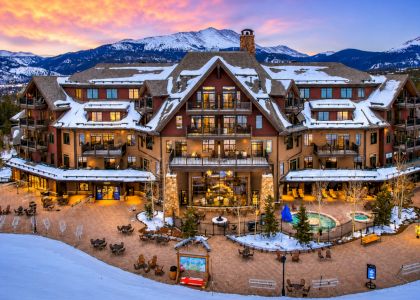 A grand ski lodge sits in a snow‑covered alpine valley, glowing warmly with lights and balconies, surrounded by mountains at sunset.