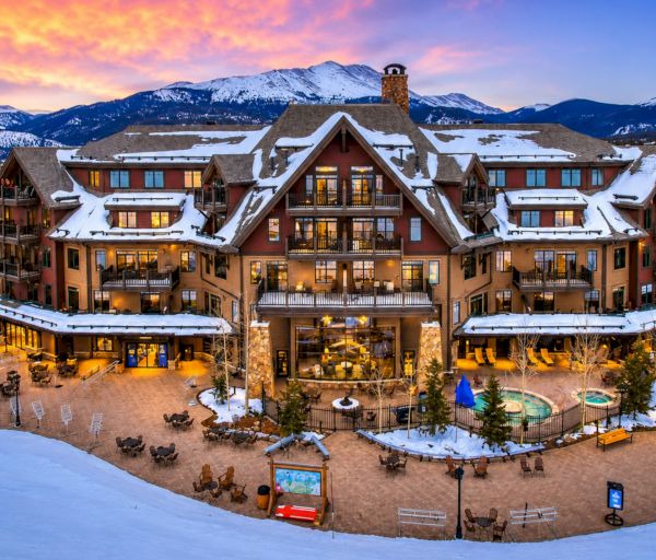 A grand ski lodge sits in a snow‑covered alpine valley, glowing warmly with lights and balconies, surrounded by mountains at sunset.