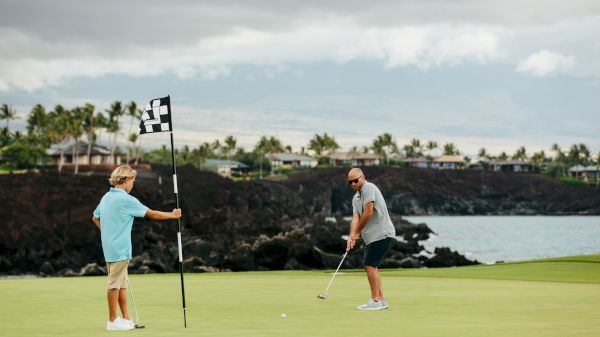 Two men play golf on a sunny course by rocky coast; one holds the flag while the other lines up a putt, palm trees in the distance.