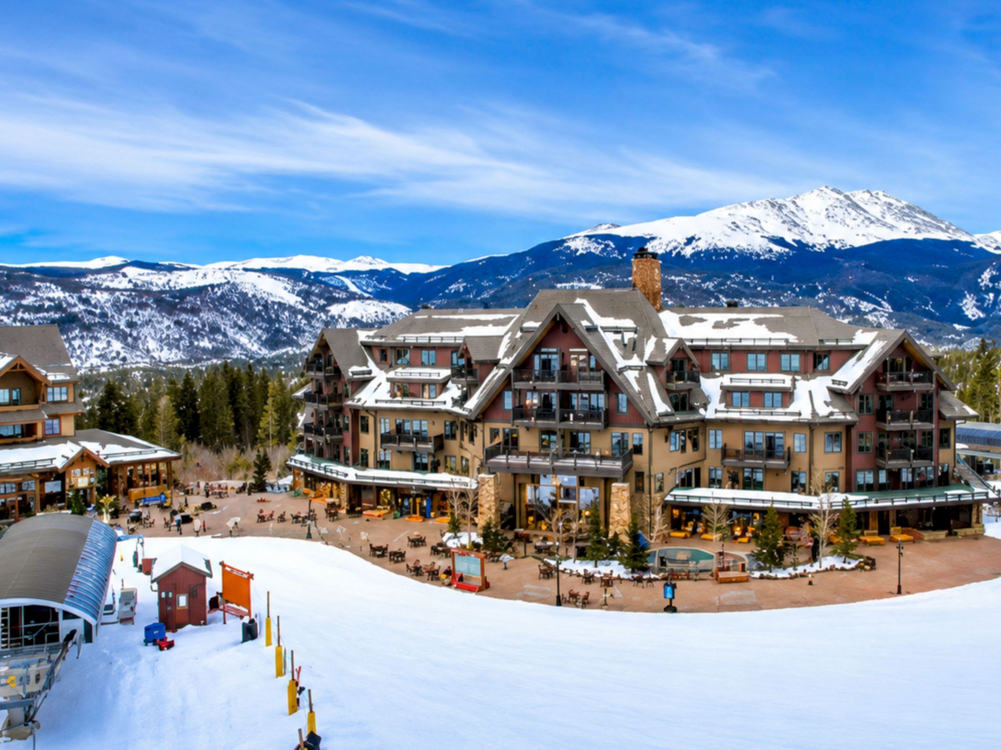 A snowy mountain village with alpine-style buildings, outdoor seating, and skiers against a backdrop of blue sky and pine trees.