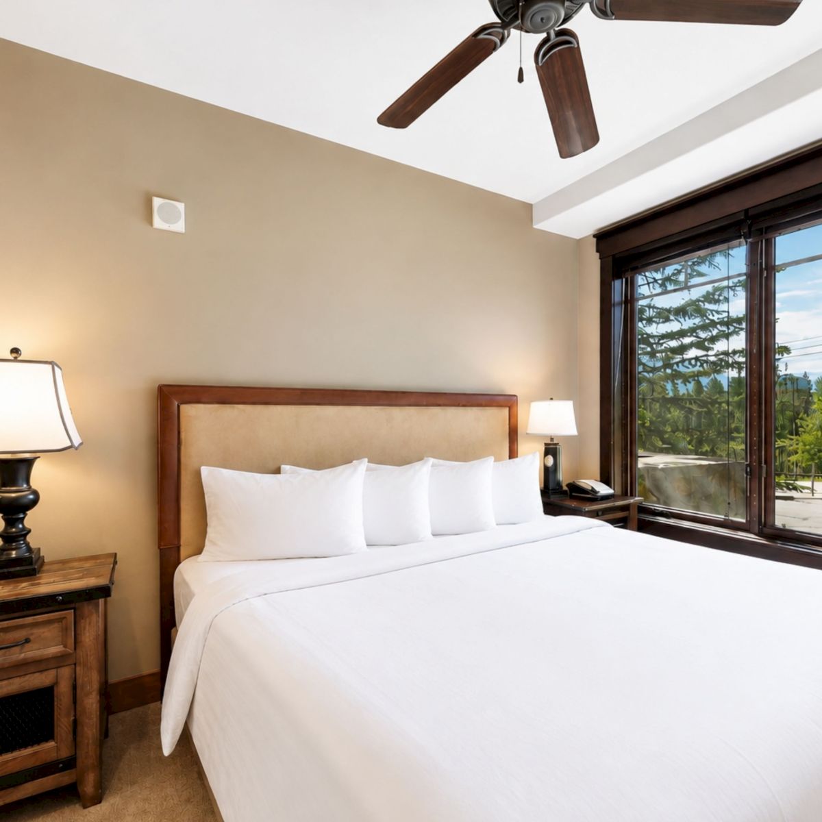 A cozy hotel bedroom with a white bed, wooden headboard, two bedside tables with lamps, a ceiling fan, and a large window overlooking trees.