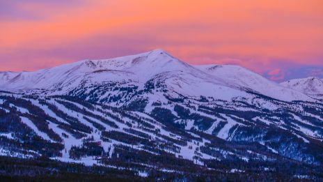 Snowy mountains with ski runs and evergreen forests beneath a pink-orange sunset sky.