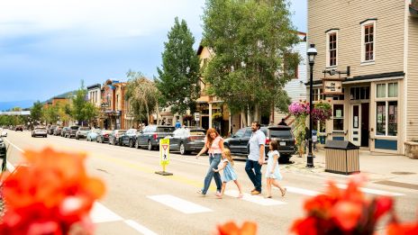 A family crossing a sunny street in a small town with shops, parked cars, trees, and colorful flowers in the foreground.