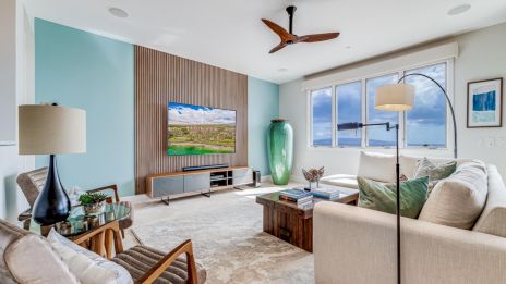 A bright, modern living room with a teal accent wall, wooden media wall, large windows, beige sofa, stylish chairs, and a ceiling fan, overlooking a scenic landscape.