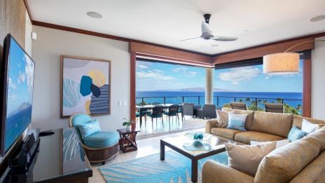 A bright coastal living room with beige sofas, a blue rug, artwork, and a wide ocean-view patio through large windows, airy and sunny.