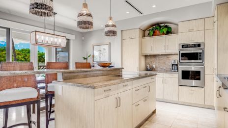 A bright, modern kitchen with light wood cabinetry, a central island, woven pendant lights, built-in ovens, and a dining area by large windows.